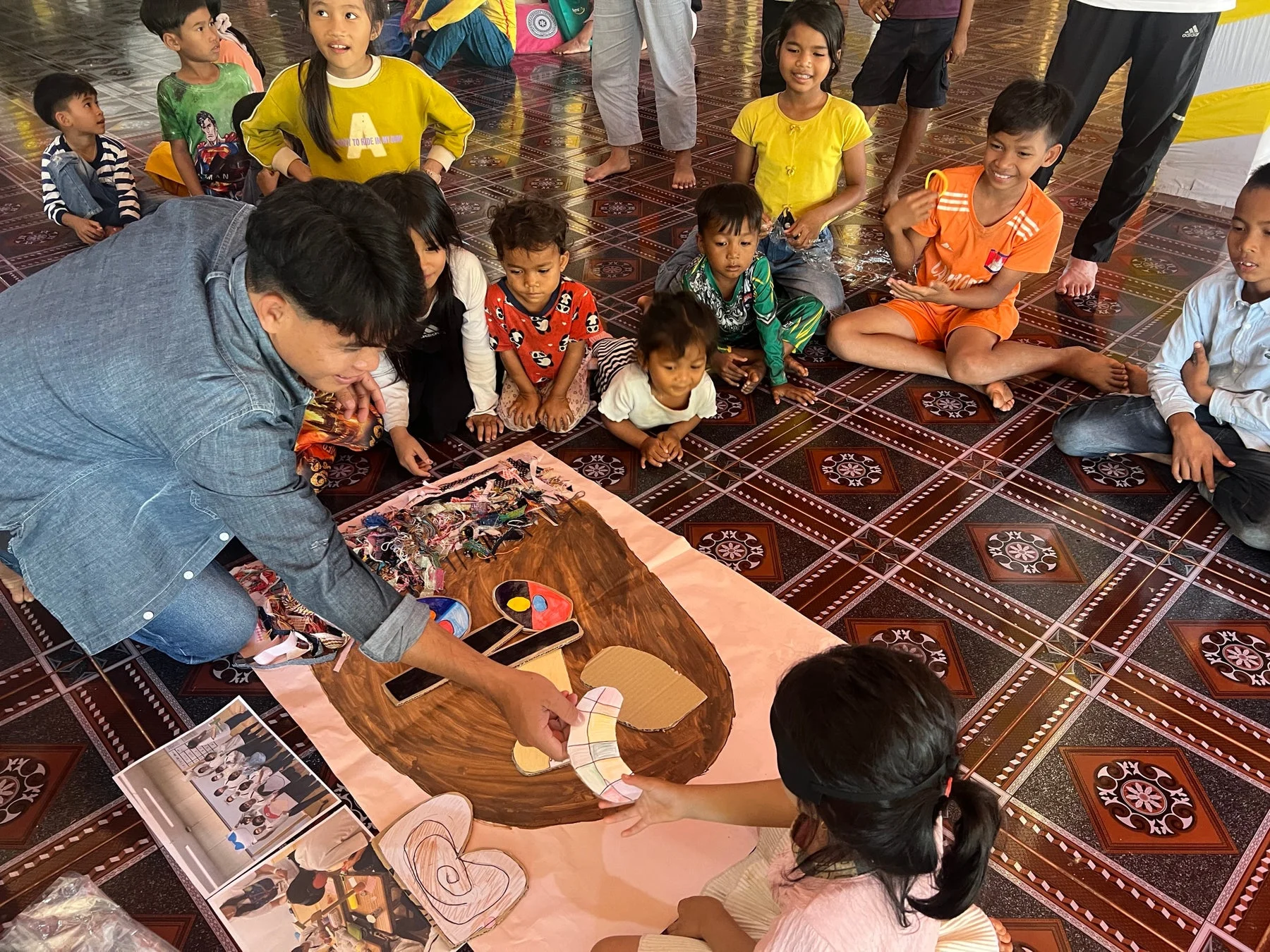 床に座った大人と子供たちが、大きな紙に絵を描いたり、段ボールや布切れを使って共同でアート作品を制作しています。