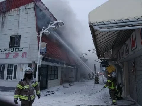 雪が積もる寒い街中で、建物から煙が激しく立ち上り、消防士たちが懸命に放水して消火活動にあたっている様子