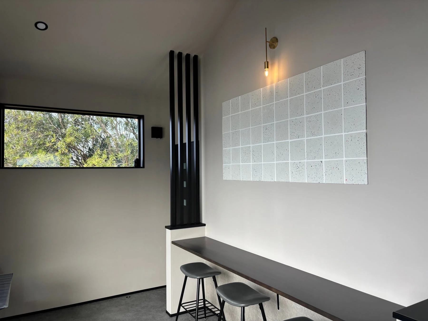 モダンでミニマリストなカフェまたはバーの店内。長いバーカウンターとバースツール、窓からは緑が見える。グリッド柄の壁アートと真鍮の壁掛け照明が特徴で、落ち着いた雰囲気の空間。