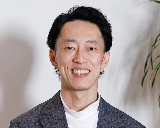 白い背景の前に立つ、白いタートルネックとダークグレーのジャケットを着た笑顔の東洋人男性のポートレート