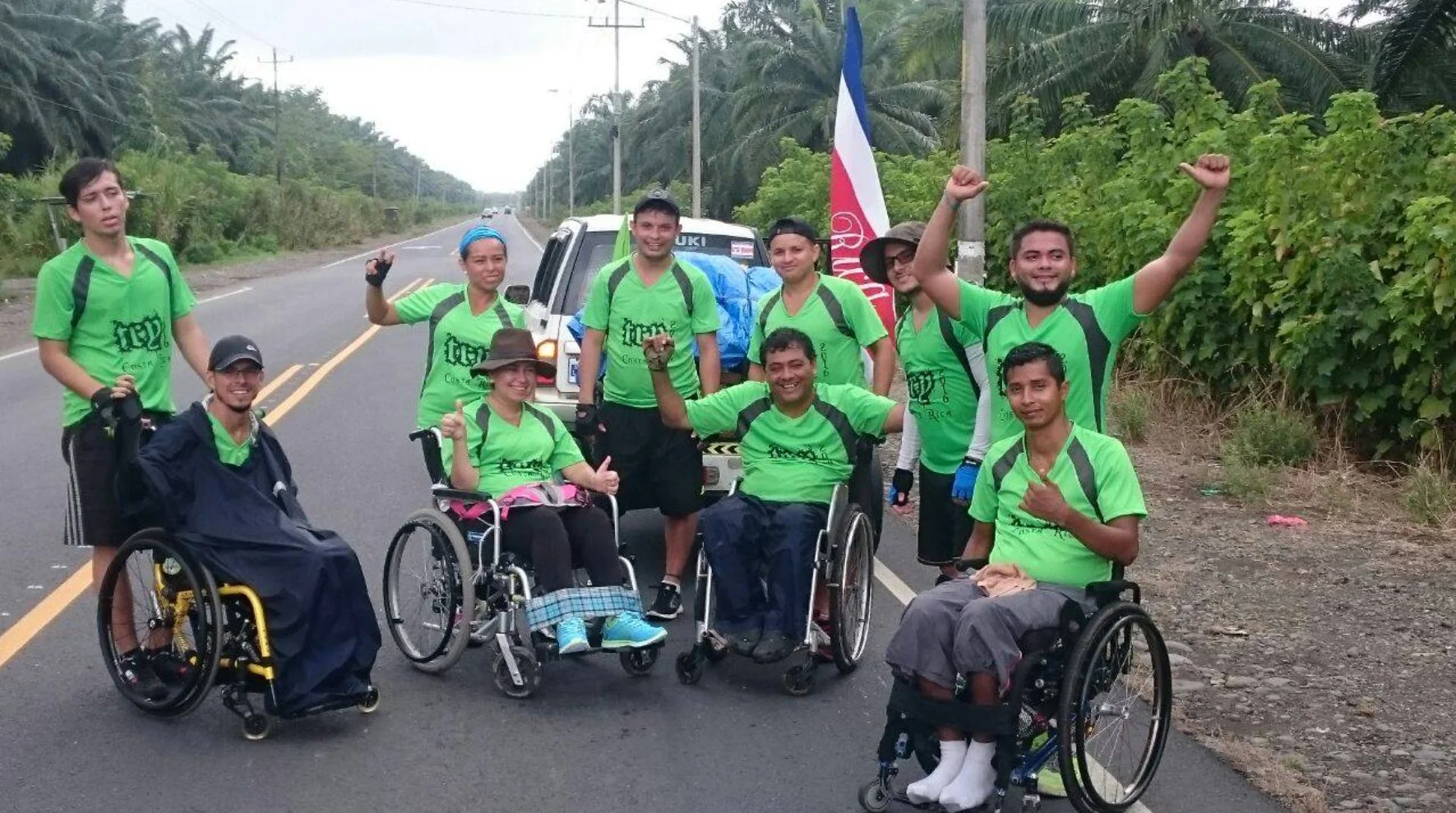緑色の揃いのTシャツを着た人々が、車椅子に乗った仲間と共に道路脇でポーズをとっています。