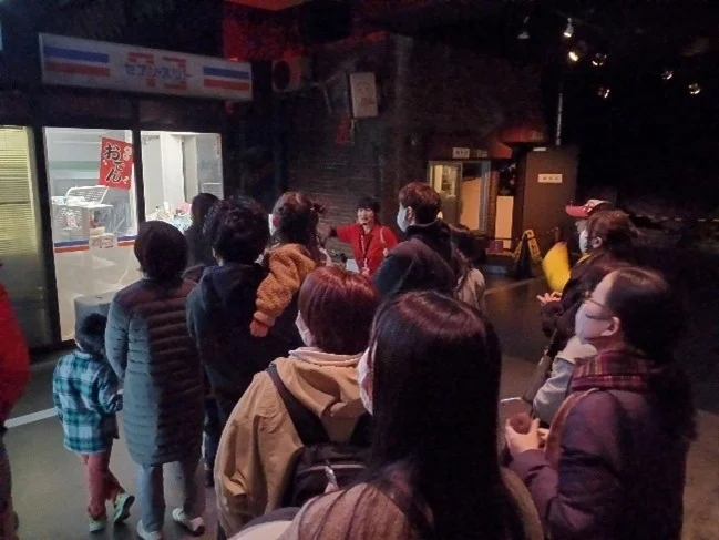 薄暗い屋内空間で、セブン-イレブン風の店舗の前に人々が列をなしている。家族連れが多く見られ、店員らしき人物が対応している。イベントやアトラクションの一環として設けられた店舗のようだ。