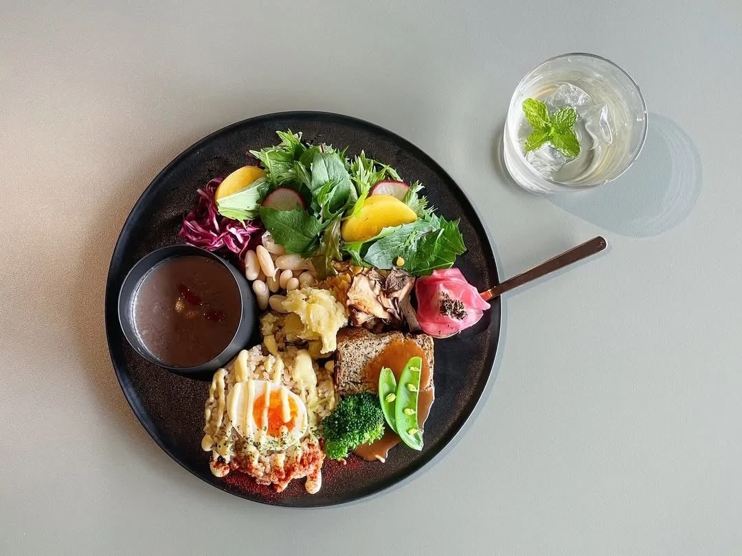 様々な種類のおかずが盛り付けられた、彩り豊かなワンプレートランチの写真です。半熟卵、パン、サラダ、スープなどがバランス良く配置されており、飲み物としてミント入りの水が添えられています。