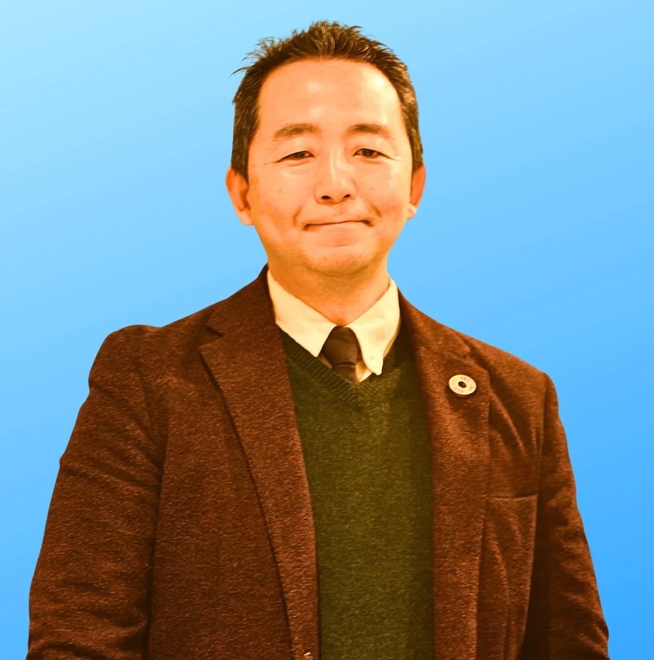 青い背景の前で、茶色のジャケットと緑のセーター、ネクタイを着用したアジア人男性が穏やかに微笑んでいるポートレート写真です。