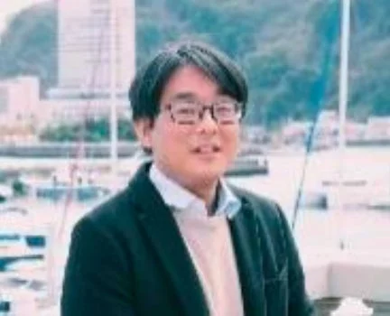 眼鏡をかけたアジア人男性が、水辺の背景を背に微笑んでいるポートレート写真です。ダークなジャケットと明るい色のトップスを着用しており、屋外で撮影された様子がうかがえます。