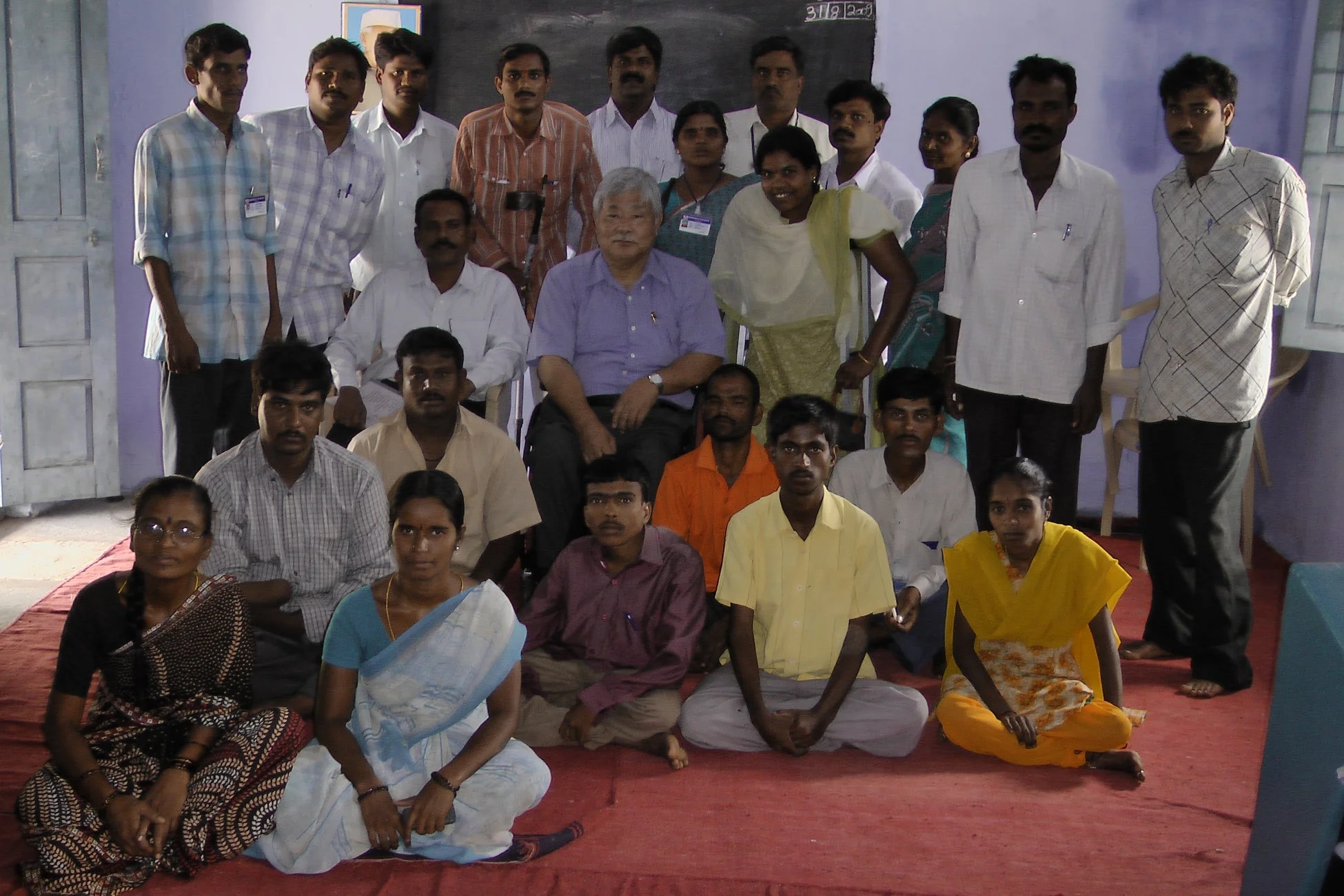 2005年3月8日、屋内の教室のような場所で、一人の東アジア人男性と多くの南アジア系の男女が集合している様子です。