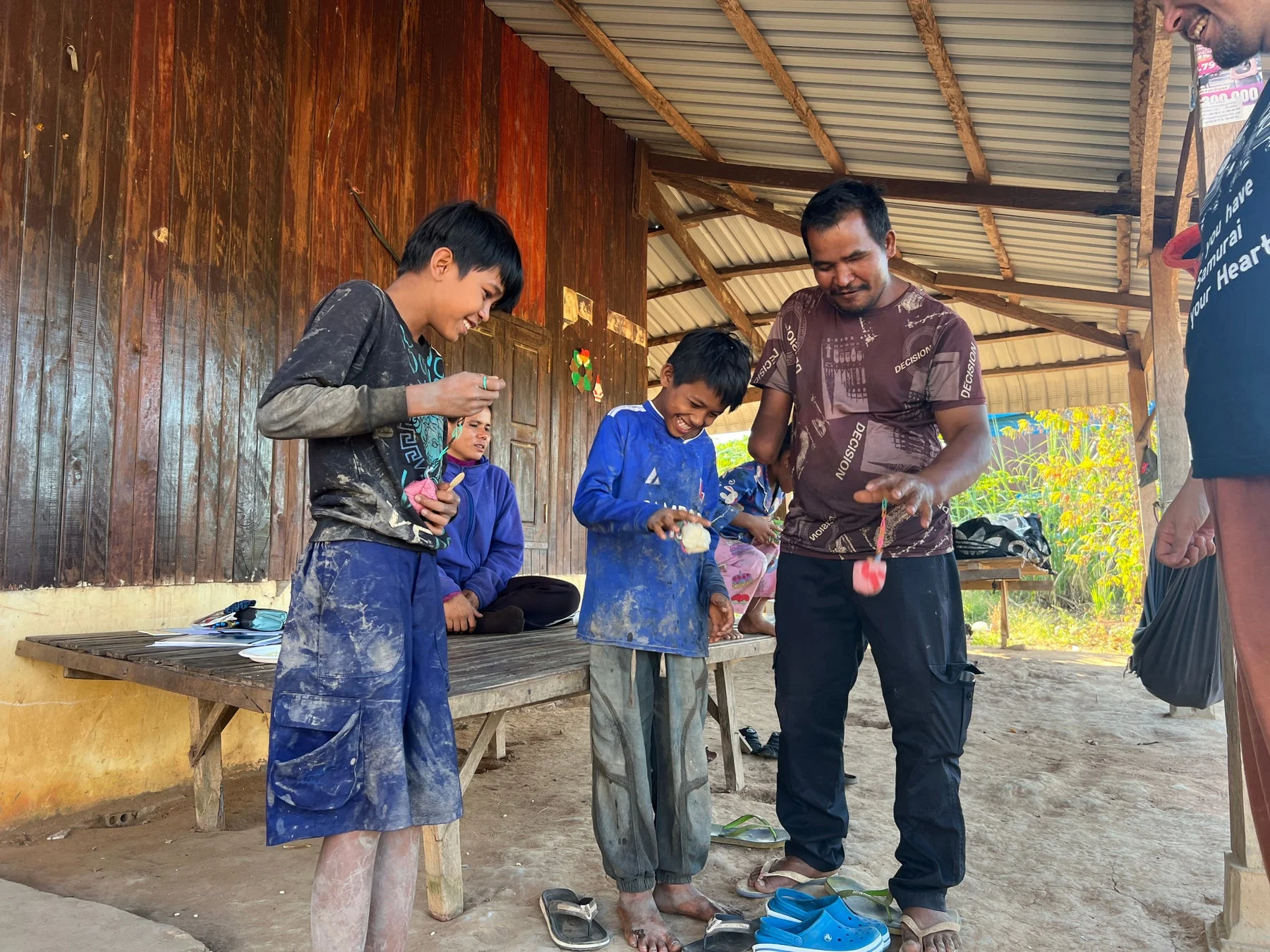 屋外の質素な家屋の前で、大人と子供たちが集まり、笑顔で交流している様子