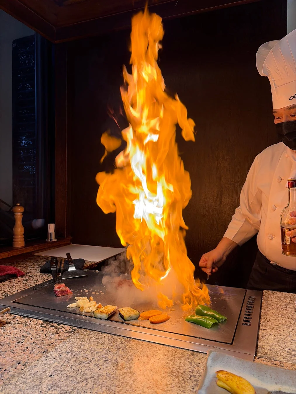 鉄板焼きのシェフが、鉄板の上で肉や野菜を調理しており、大きな炎が上がっているダイナミックな瞬間を捉えた画像です。