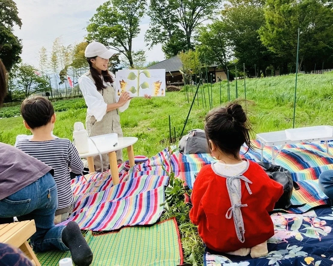広々とした屋外の芝生の上で、一人の女性が子供たちに絵本の読み聞かせをしている場面