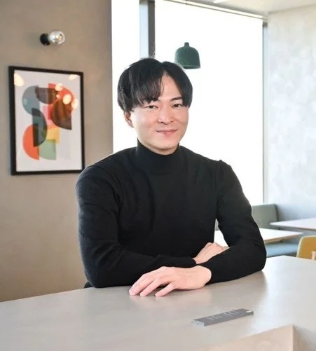 黒いタートルネックを着たアジア人男性が、モダンな雰囲気の屋内でテーブルに腕を置いて座り、穏やかな笑顔でカメラを見ています。背景には抽象的なアートが飾られています。