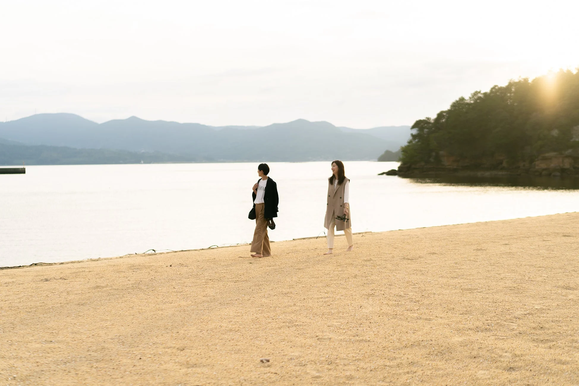 砂浜を裸足で歩く二人の女性が写っている。背景には海と山が見え、夕日が差し込む穏やかな風景で、リラックスした雰囲気を感じさせます。