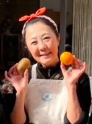 赤いリボンのカチューシャとエプロンを着用した女性が、笑顔でじゃがいもとみかんをそれぞれ手に持っている様子です。