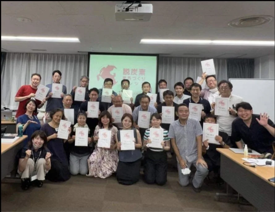 会議室で多くの参加者が修了証のような書類を手に笑顔で写っている集合写真です。背景のスクリーンには「脱炭素 暮らしづくり」と表示されており、環境に関するセミナーや研修の終了記念撮影と推測されます。