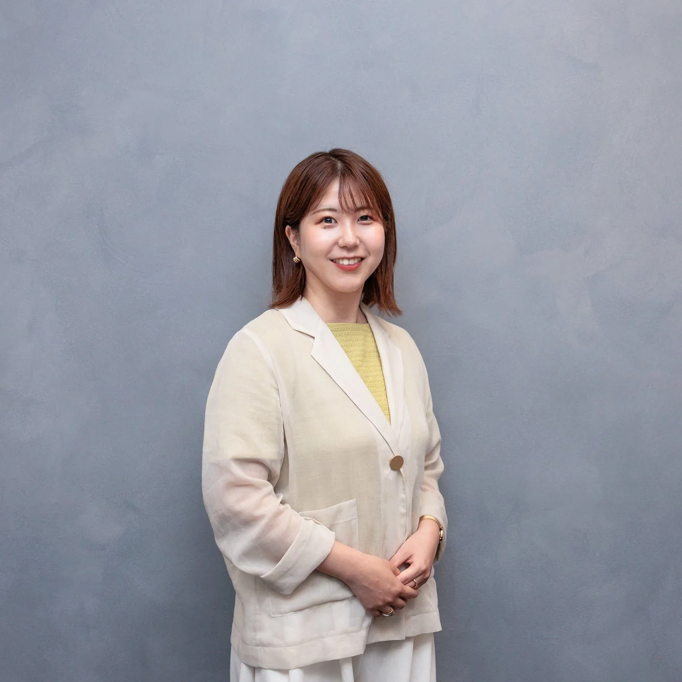 グレーの壁を背景に、明るい色のジャケットと黄色のトップスを着用した笑顔の女性のポートレートです。