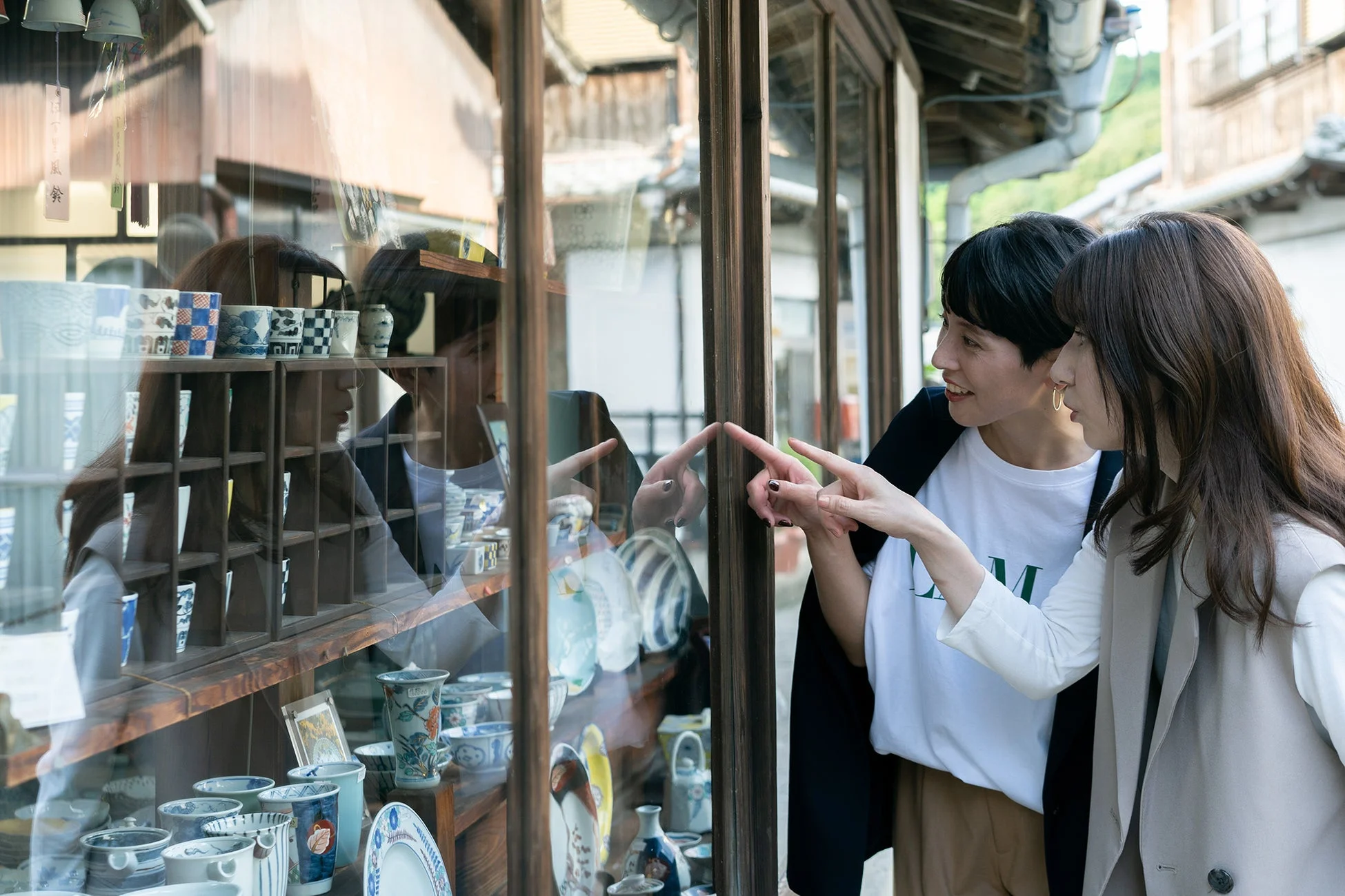 二人の女性が伝統的な雰囲気の店先で、窓越しに並べられた陶器や食器を眺めています。楽しそうに指を差し、会話している様子がうかがえ、和やかなひとときを過ごしているようです。