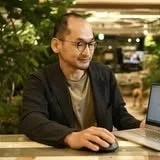 眼鏡をかけた男性が、緑のある屋内の環境でノートパソコンとマウスを使って作業に集中している様子。ビジネスシーンやリモートワークを連想させます。