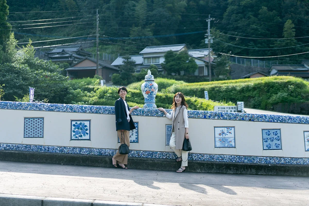 笑顔の女性二人が、有田焼のような青と白の陶磁器で装飾された壁の前に立っています。背景には日本の伝統的な家屋と豊かな緑の山々が広がっています。