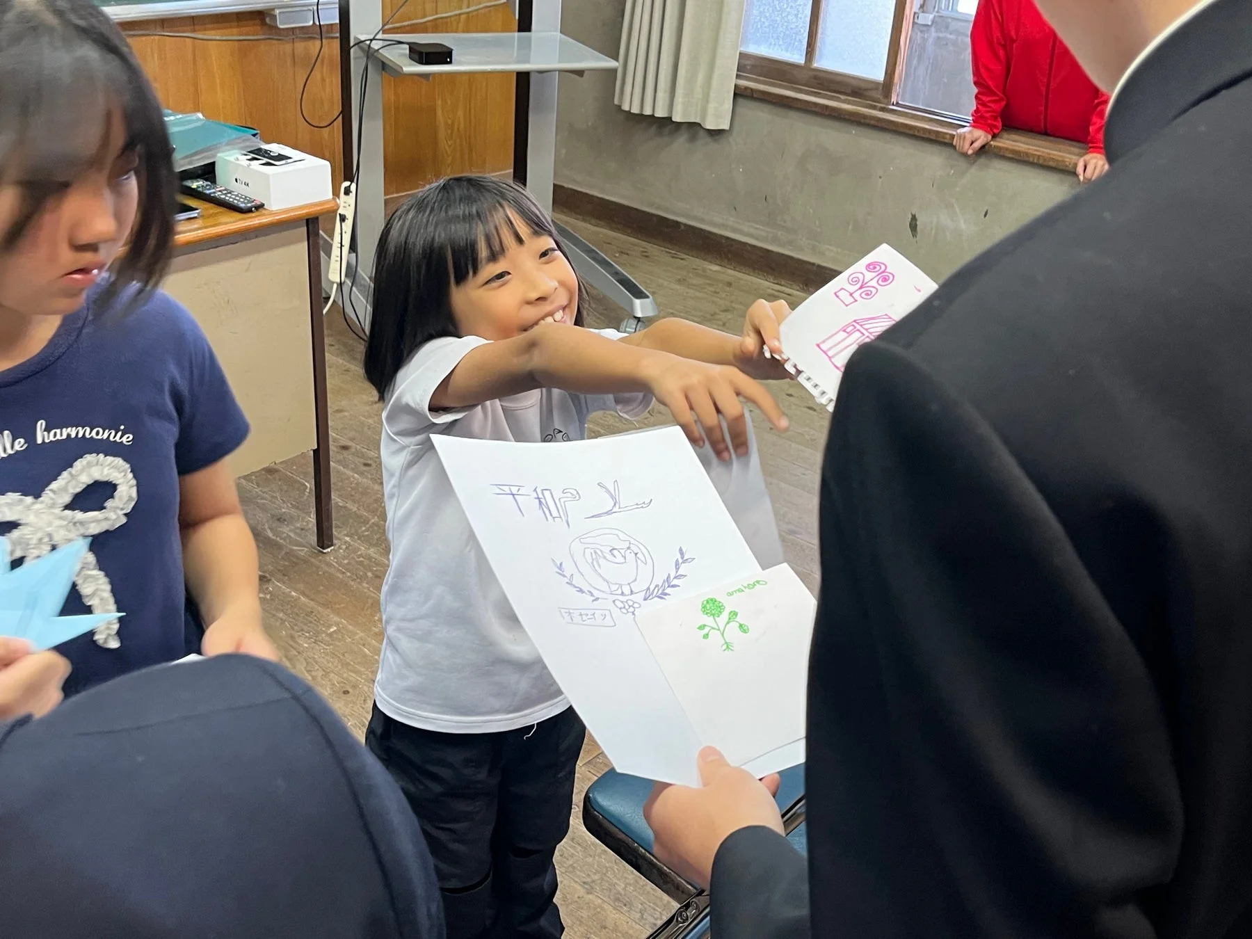 教室らしき場所で、笑顔の子供が「平和」と書かれた絵を大人に見せている様子
