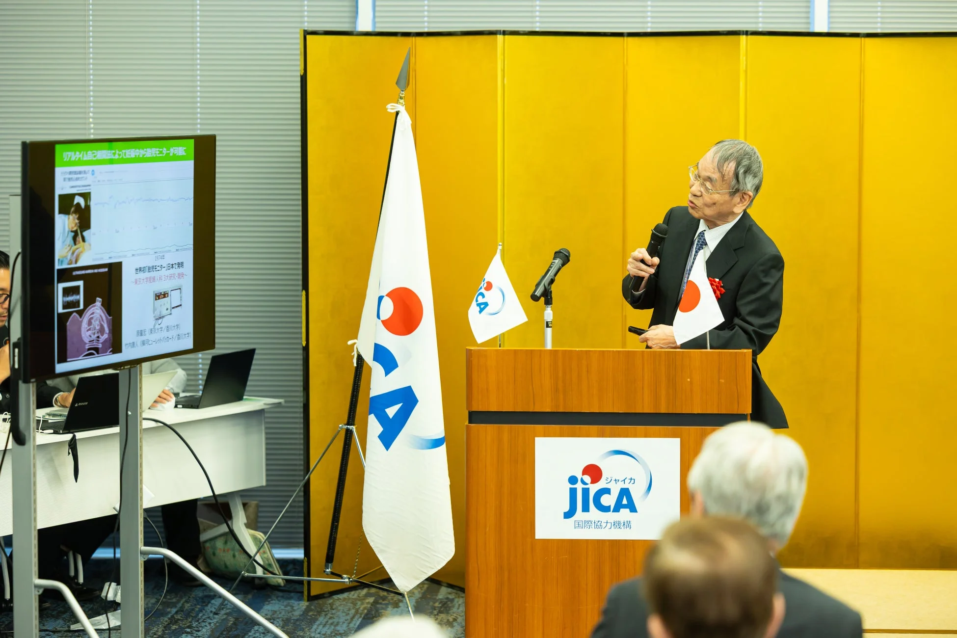 JICAのイベントで、高齢の男性が演台に立ち、マイクと日本の国旗を持ちながらプレゼンテーションを行っています。