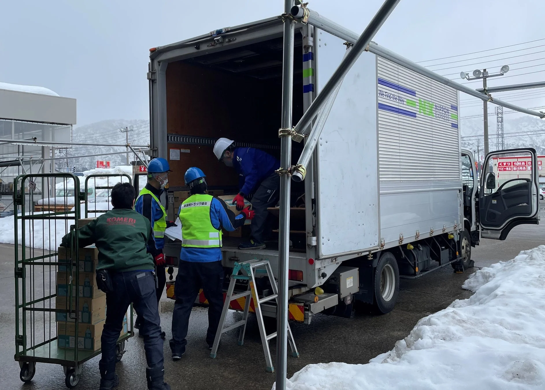 雪が積もる屋外で、日本通運のトラックに複数の作業員が荷物を積み込んでいる。コメリのスタッフや新潟県トラック協会のベストを着た人々が天然水の箱を運んでおり、冬場の物流や支援活動の様子がうかがえる。