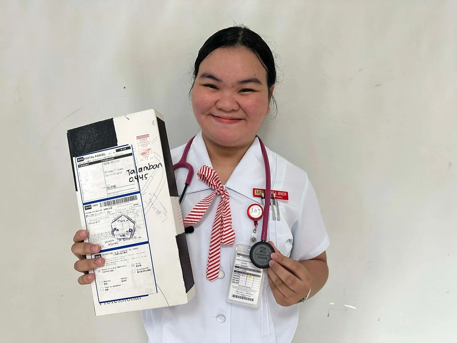 看護師か看護学生と思われる制服姿の若い女性が笑顔で小包と聴診器を持っている写真です。