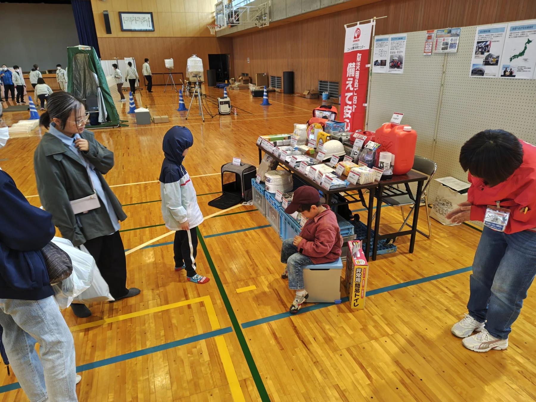 体育館で行われている防災イベントの様子。コメリ災害対策センターのブースで、非常用簡易トイレや様々な防災用品が展示され、親子連れが関心を持って見学している。