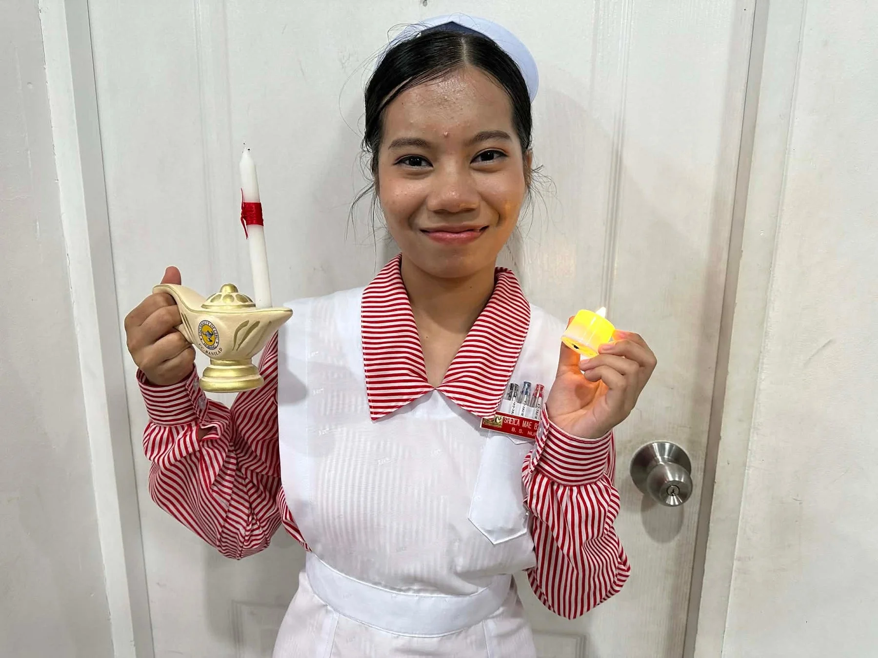 看護師の制服を着た女性が、ナイチンゲールランプとLEDキャンドルを持って笑顔で立っています。