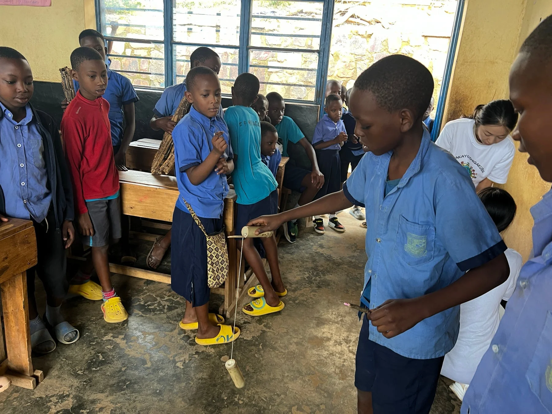 教室で多くの子どもたちが集まり、一人の男の子が手作りのようなおもちゃで遊んでいる様子