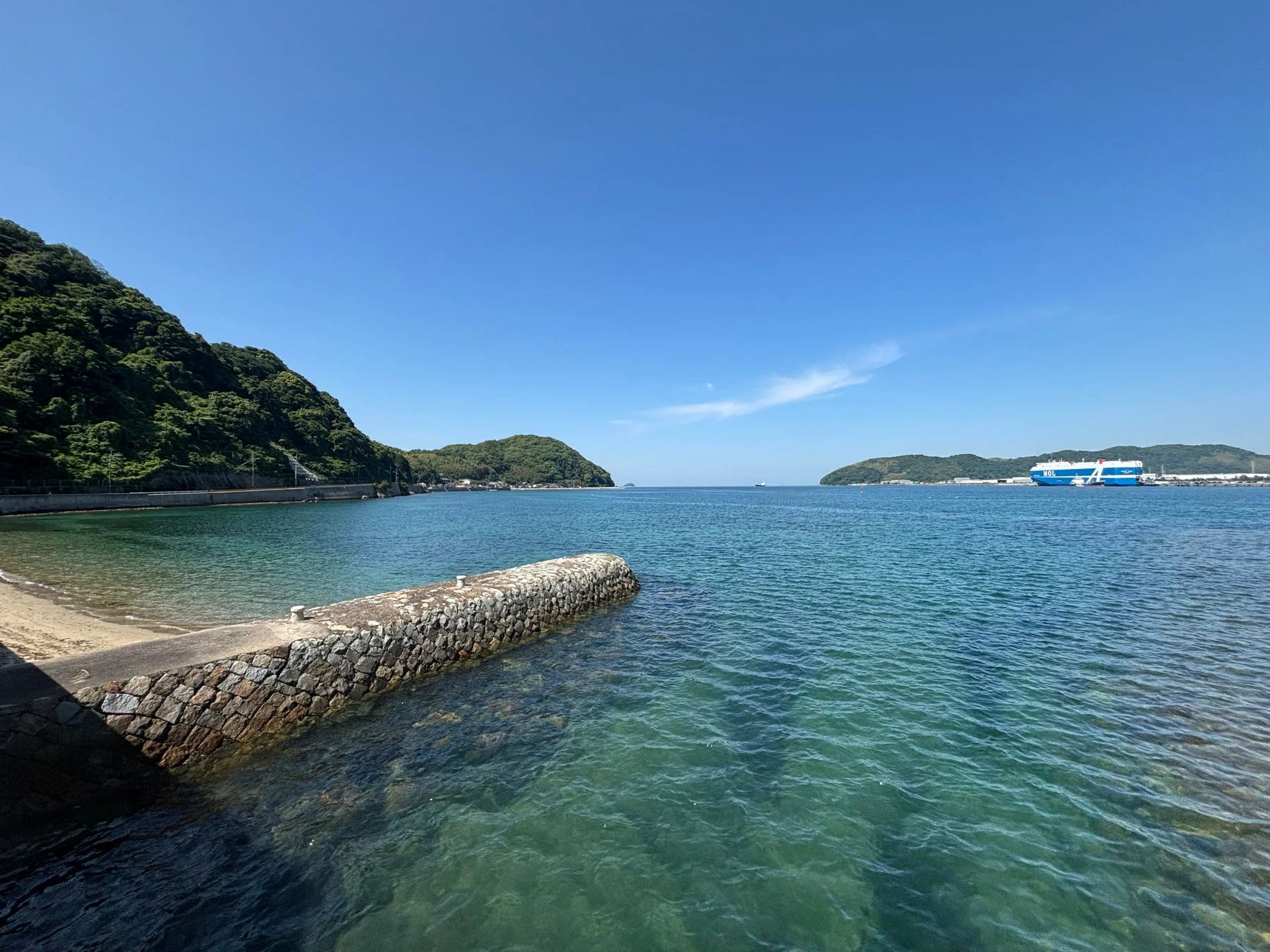 晴れた日に広がる青い空と穏やかな海、緑豊かな山々が美しい海岸風景です。遠くには大きな青い船が停泊しており、手前には石積みの堤防が見えます。