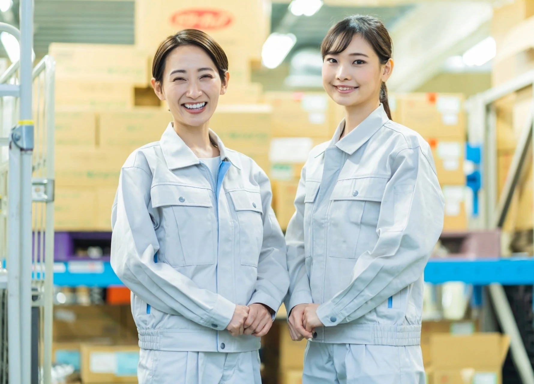 倉庫のような場所で、グレーの作業服を着た2人の女性が笑顔で並んでいます。明るくプロフェッショナルな雰囲気です。