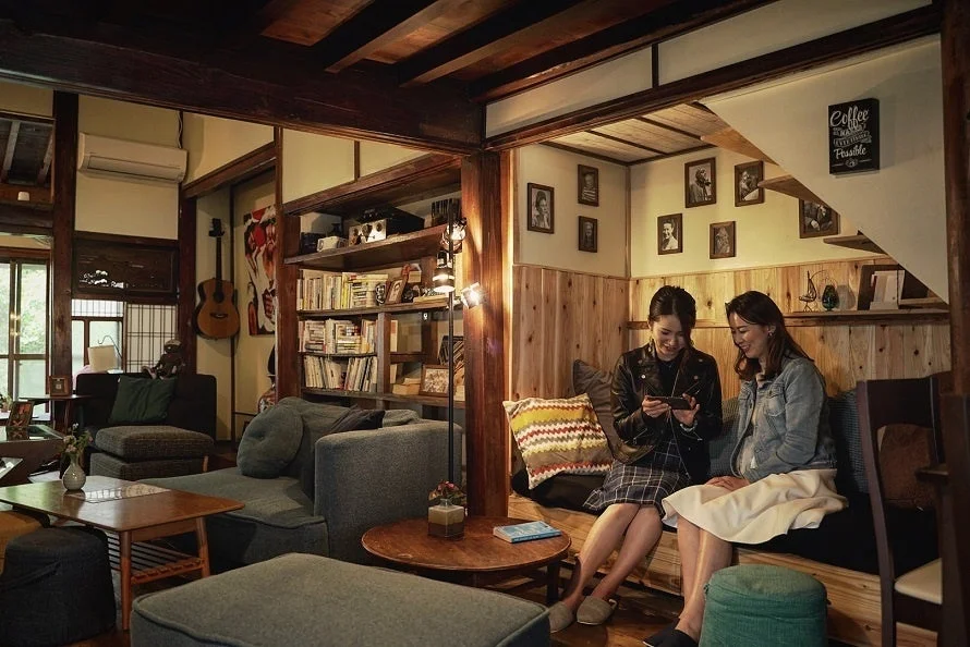 古民家風のカフェで、二人の女性がソファに座り、スマートフォンを見ながら楽しそうに会話している様子。温かみのある照明と木製のインテリアが落ち着いた雰囲気を醸し出しています。