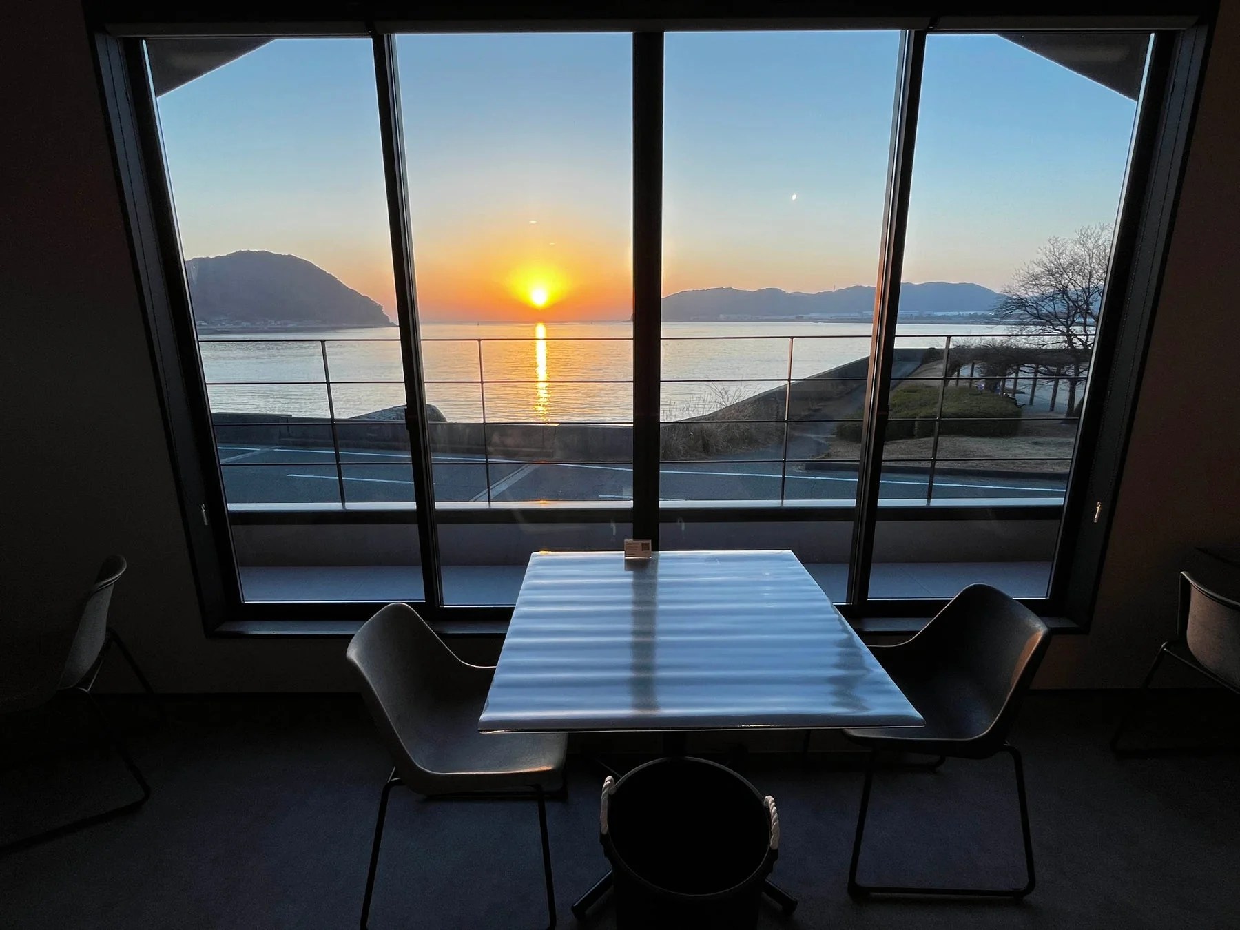 窓から見える美しい夕焼けの海の景色です。室内にはテーブルと椅子があり、沈む夕日が海面を明るく照らしています。遠景には山々が広がり、穏やかで開放的な雰囲気が感じられます。