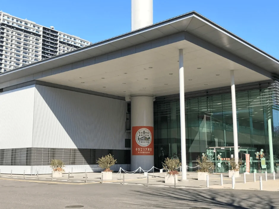 青空の下、現代的なデザインの商業施設が写っています。ガラス張りの外観と白い柱が特徴的で、手前には植木が配置されています。背景には高層マンションが見えます。