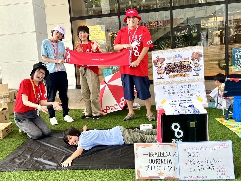 「一般社団法人80億RITAプロジェクト」の屋外イベントで、人々がビニール袋で三角巾を作る方法を学んでいます。災害時の健康法やミニ手話講座も提供され、防災意識を高めるための教育的な催しです。