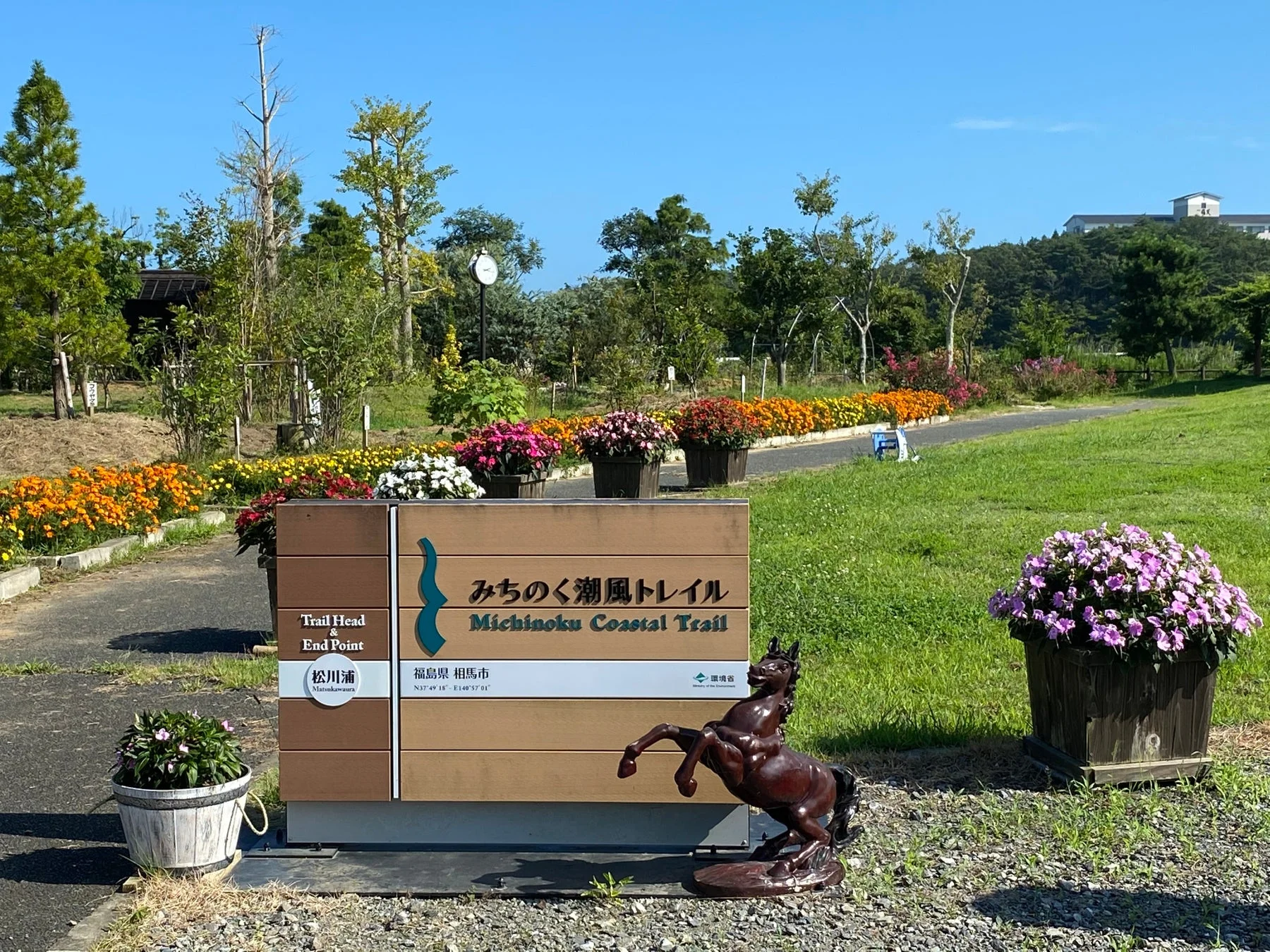 福島県相馬市の松川浦にある「みちのく潮風トレイル」の案内板が写っています。色とりどりの花壇や芝生が広がり、青空の下、馬の像がアクセントとなっています。トレイルの出発点・終点を示す標識です。