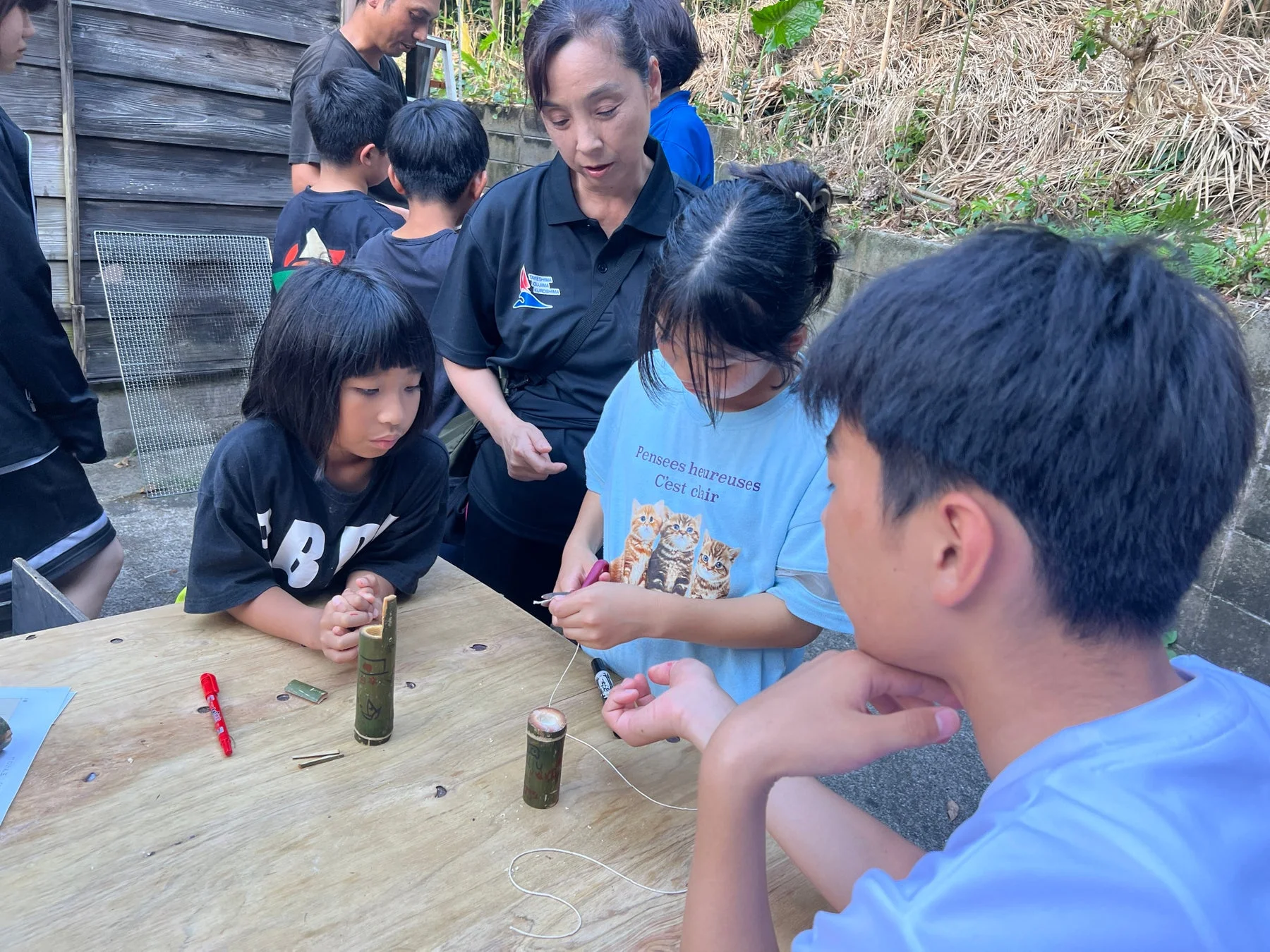 屋外の木製テーブルで、子供たちが大人の指導のもと竹細工のようなクラフト活動に集中している様子