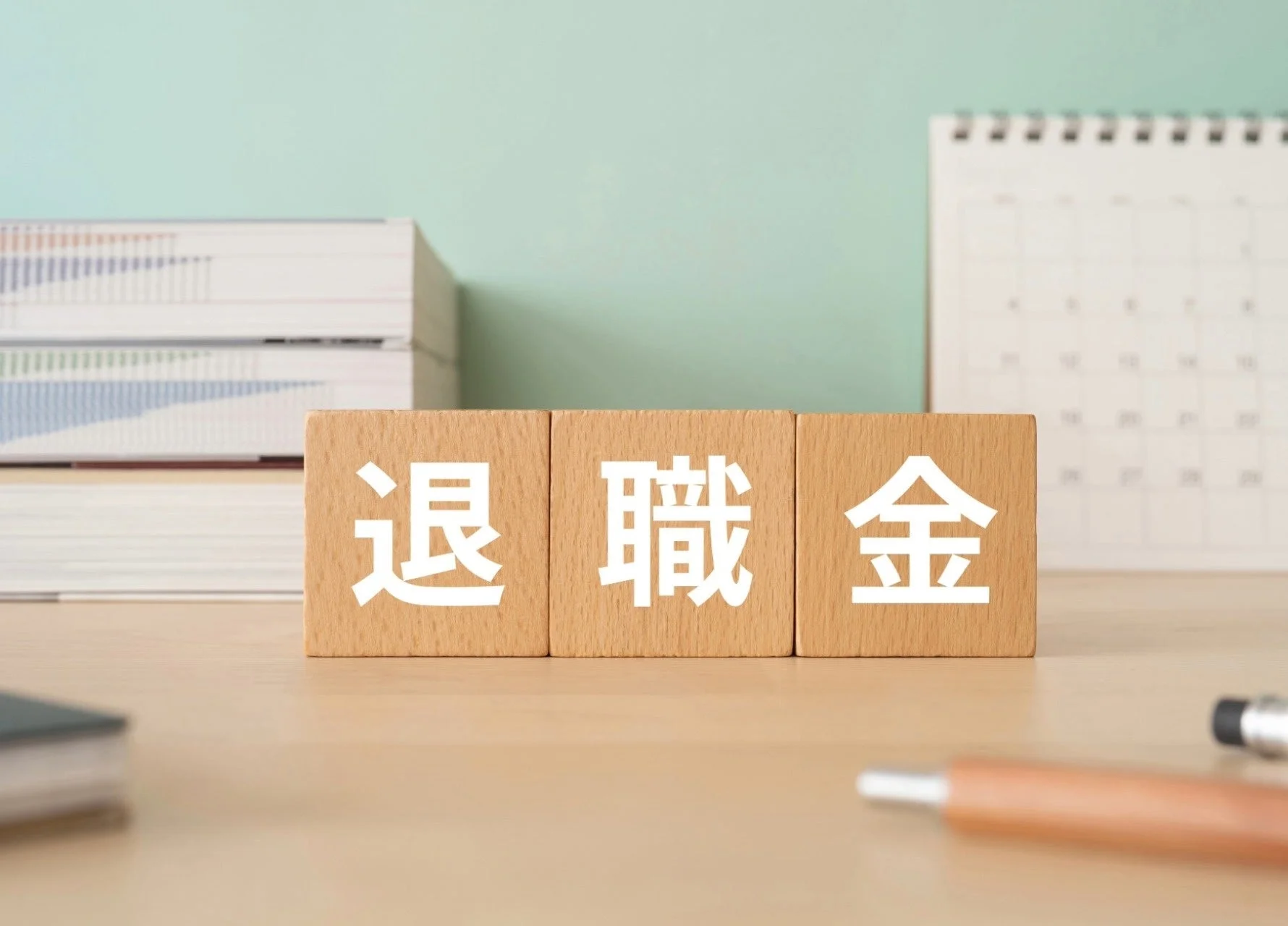 「退職金」と書かれた木製のブロックが机の上に並べられています。背景には書類の束やカレンダーがあり、退職後のライフプランや資産形成について考えさせるイメージです。