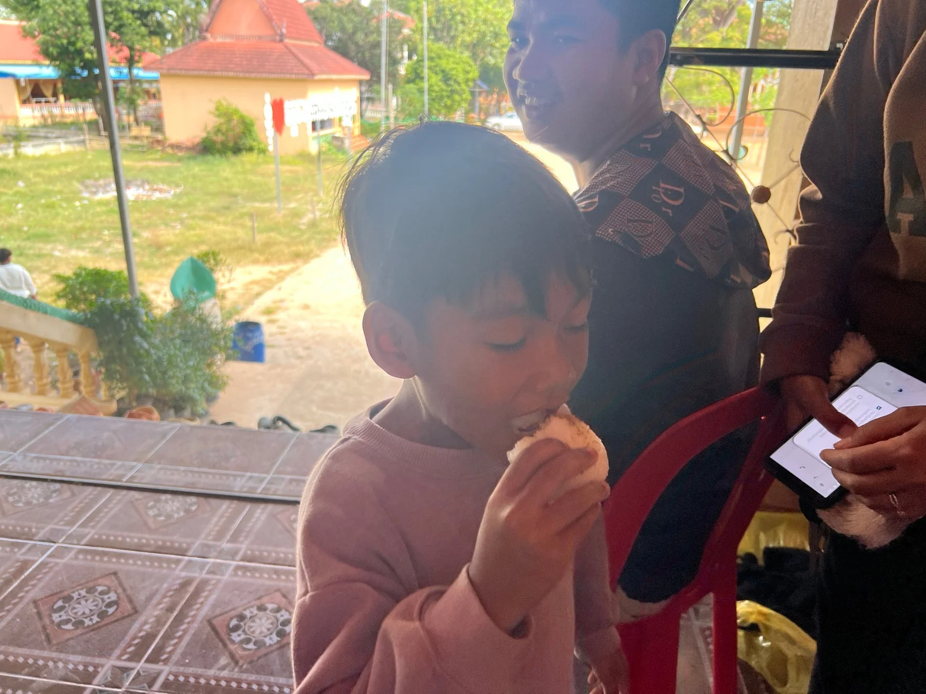 屋外で少年が白い食べ物を口に運んでおり、背景には笑顔の男性とアジア風の建物が見える