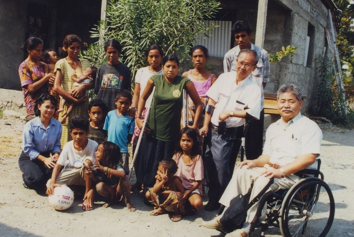 開発途上国の地域で、地元住民と日本人男性2名（うち1名は車椅子）が屋外で集合している写真。