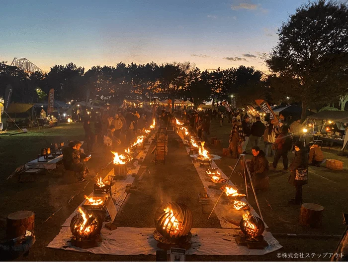 夕暮れ時の屋外イベントで、多くの人々が焚き火を囲んで集まっています。暖かな炎が会場を照らし、賑やかで心地よい雰囲気が広がっています。