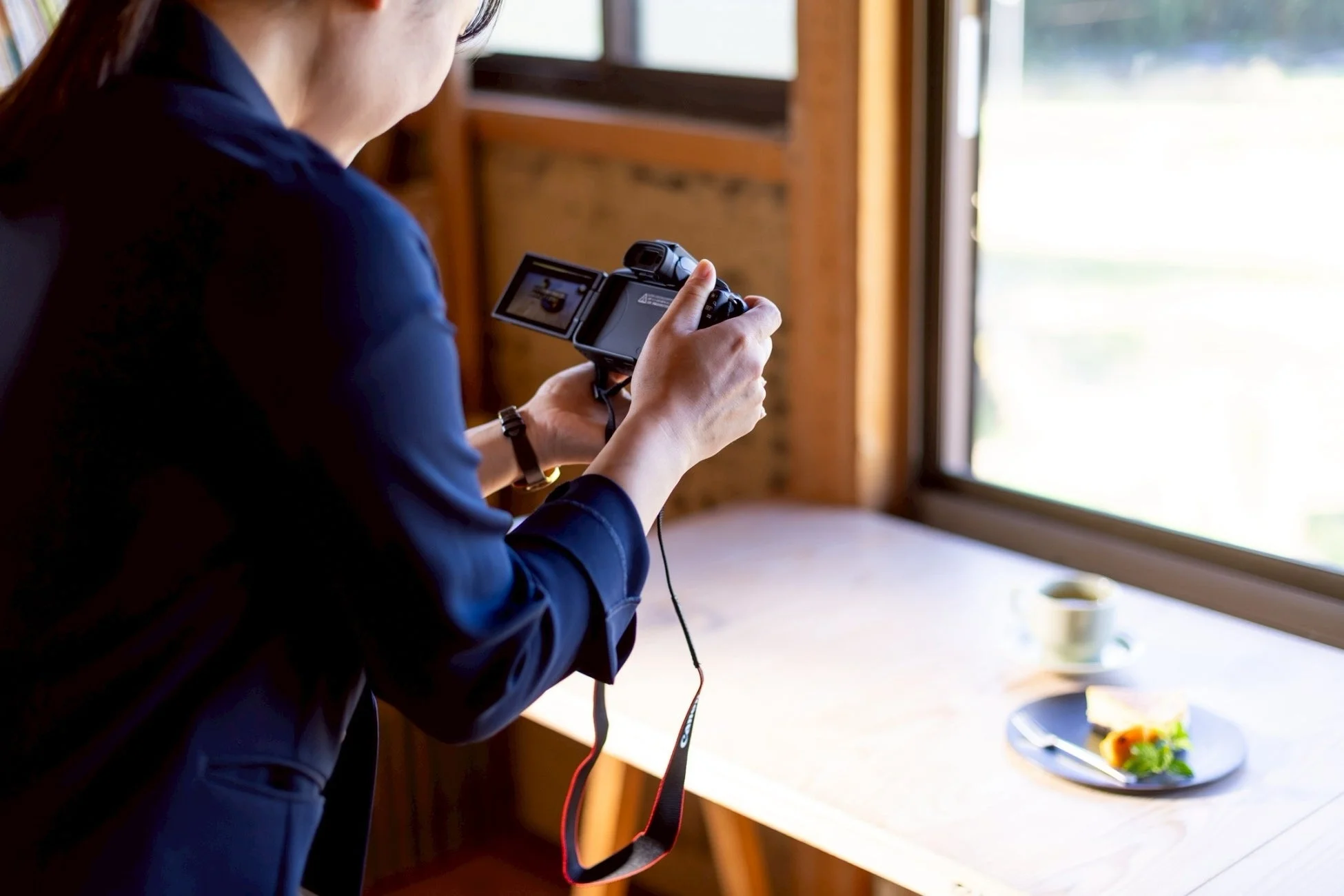 窓際の木製テーブルに置かれたケーキとコーヒーを、デジタルカメラのバリアングル液晶画面を見ながら撮影している人物の後ろ姿