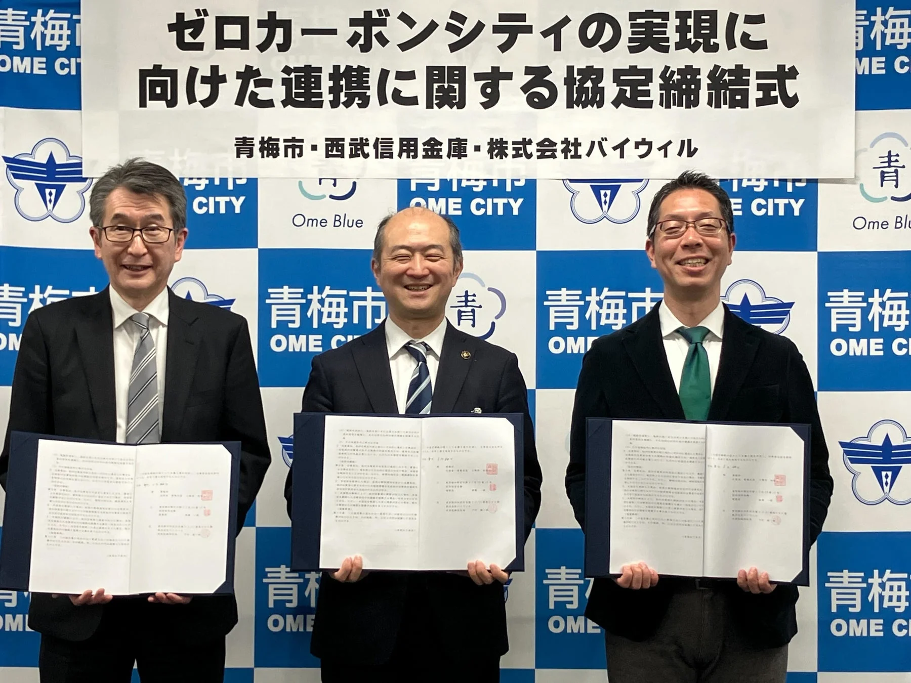 青梅市、西武信用金庫、株式会社バイウィルの3者がゼロカーボンシティ実現に向けた連携協定を締結している様子。3名の代表者が協定書を手に笑顔で並んでいる。