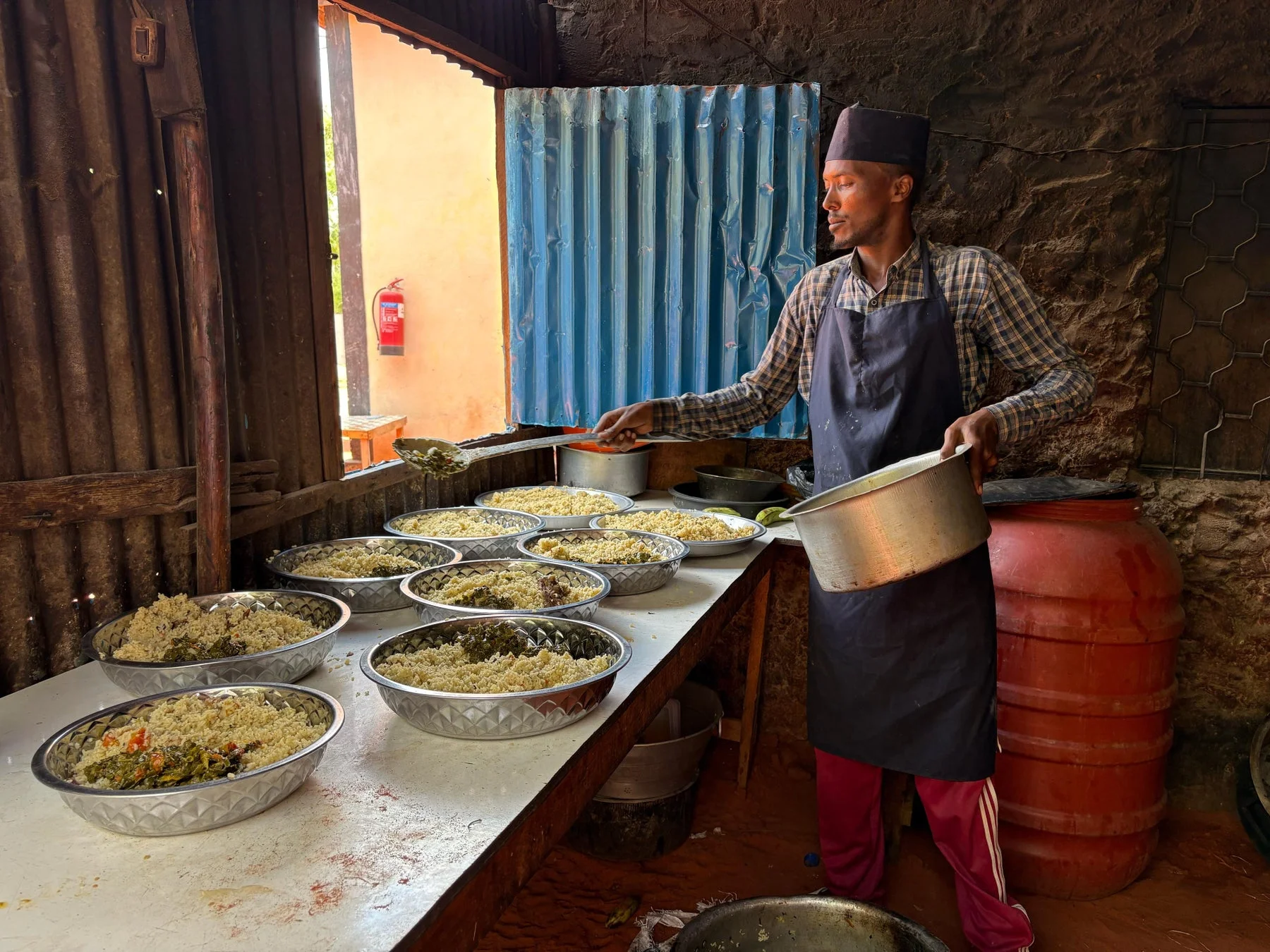 エプロンと帽子を着用した男性が、多くの金属製ボウルに盛られた米やクスクスのような料理を準備している様子です。