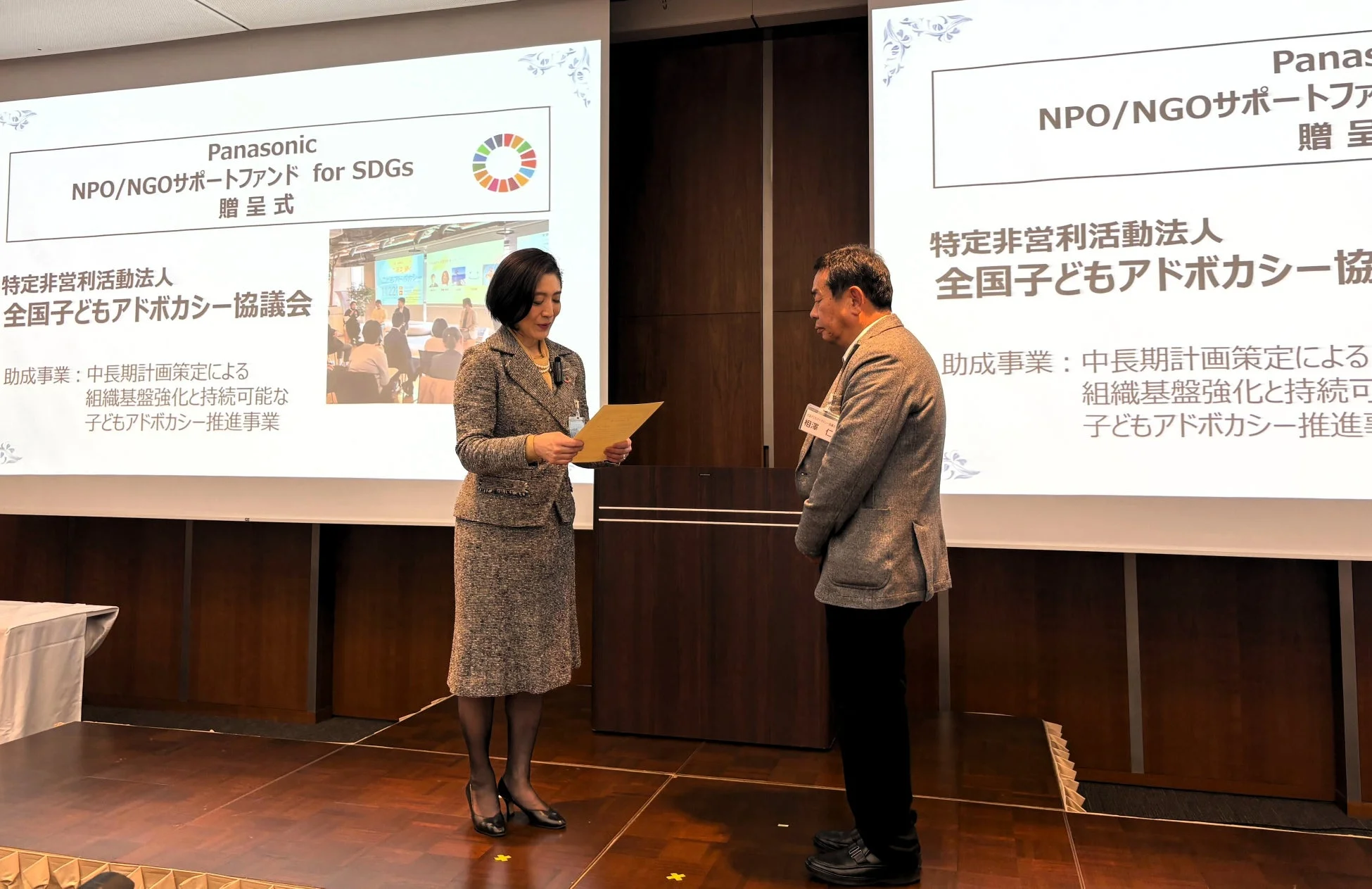 パナソニックによるSDGs NPO/NGOサポートファンドの贈呈式の様子。女性が男性に書類を手渡し、全国子どもアドボカシー協議会への助成がスクリーンに表示されている。