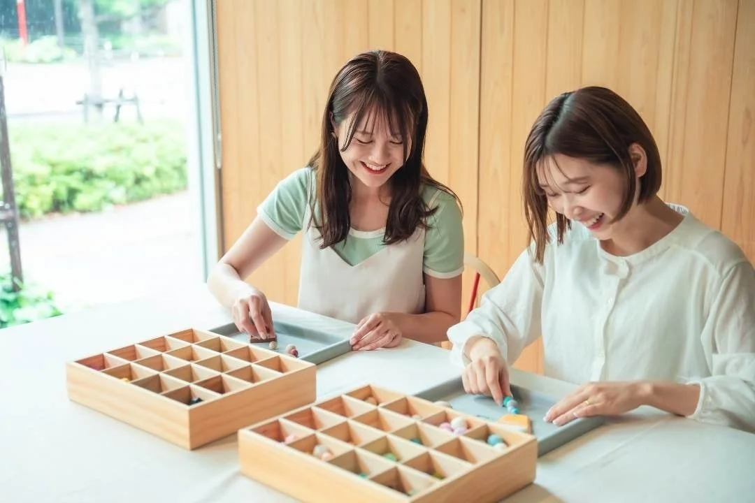 室内で2人の若い女性が笑顔で、小さなカラフルなアイテムを木箱に仕分けたり、手作りの作業に楽しそうに取り組んでいる様子