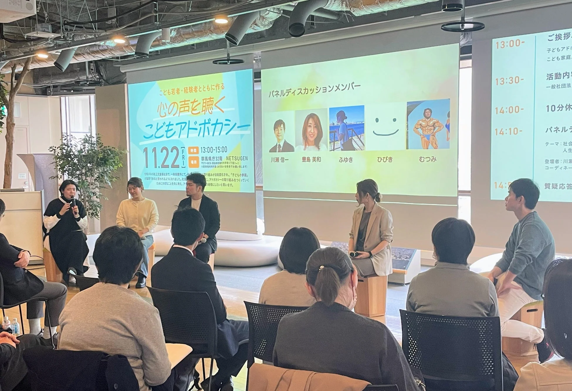 「こどもアドボカシー」に関するイベント会場の様子。スクリーンにはイベント詳細やパネルディスカッションメンバーが表示され、登壇者たちが聴衆に向けて話をしている。多くの参加者が集まり、熱心に耳を傾けている。