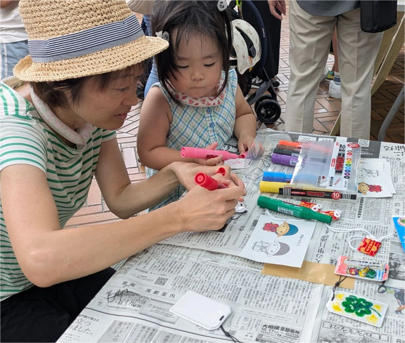 麦わら帽子をかぶった女性と小さな女の子が、新聞紙を敷いたテーブルで色鉛筆を使ってクラフト活動を楽しんでいる様子
