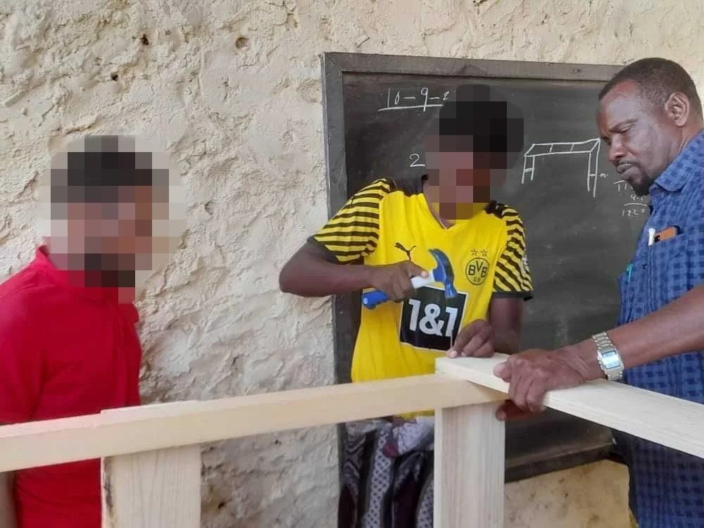 大人が若者に木工を指導している様子。若者はハンマーで木材を組み立て、黒板には図面が描かれている。職業訓練や実習の場面で、学習と実践が行われている。