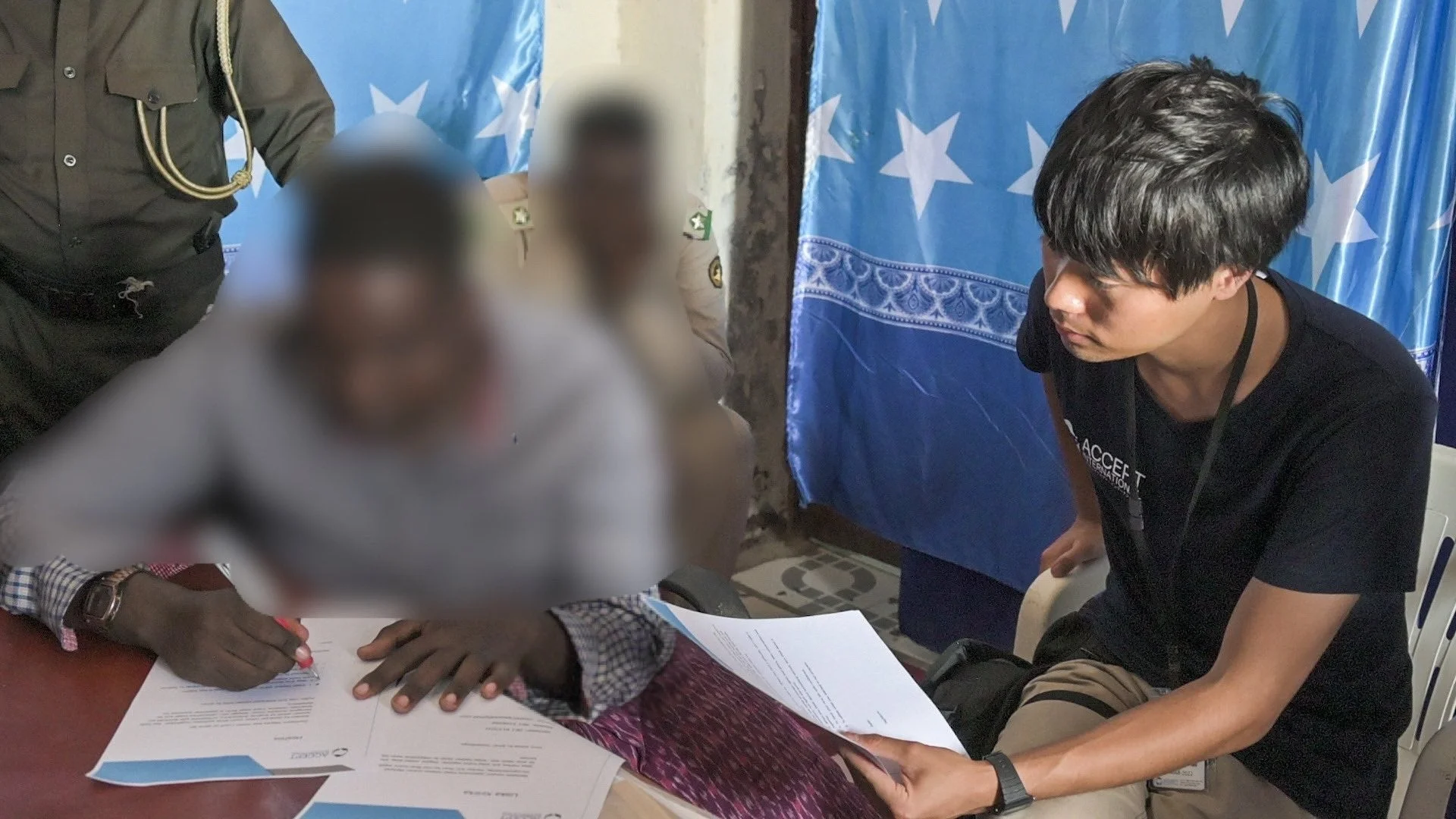 ソマリア国旗が背景にある室内で、一人の男性が書類に署名しています。その傍らには「ACCEPT INTERNATIONAL」と書かれたTシャツを着た若い男性がおり、協力関係の事務作業を行っている様子がうかがえます。