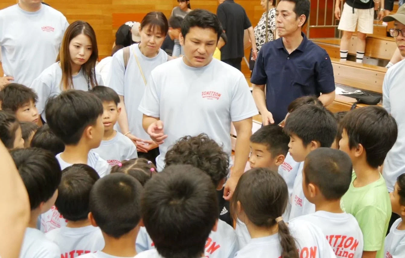 体育館で、多くの子供たちが一人の男性を囲んで話を聞いている様子